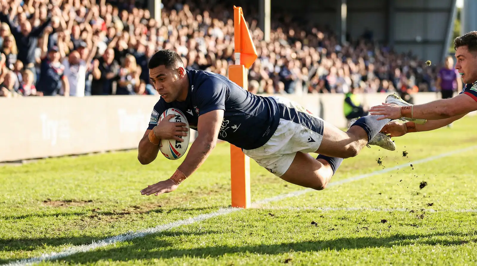 Rugby league winger diving to score a try in the corner of a grass pitch under floodlights